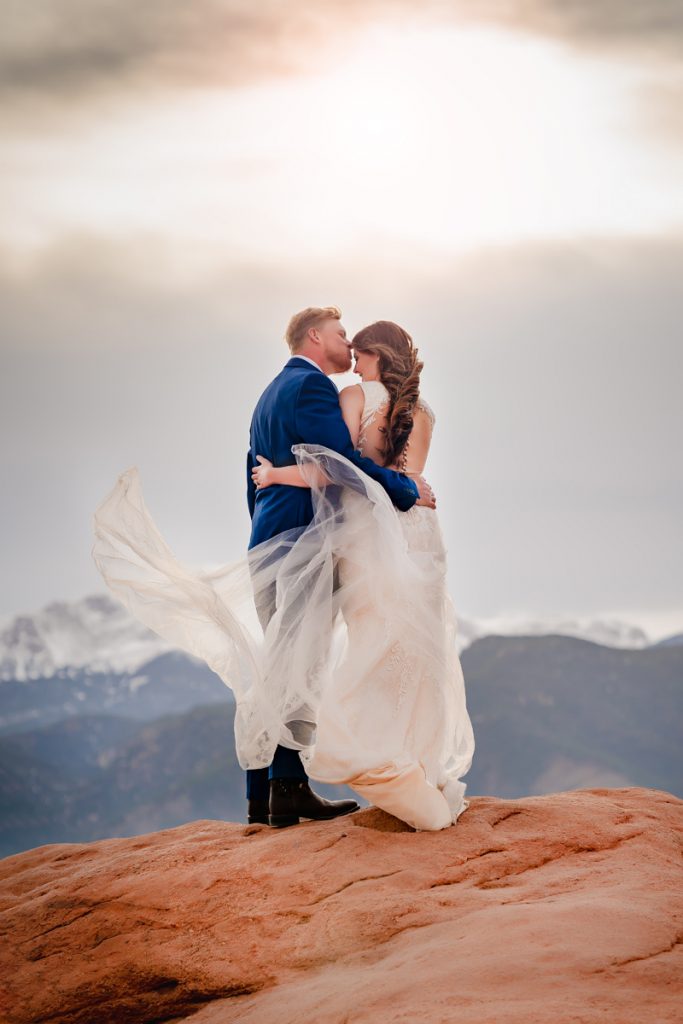 Elopement Photography, A bride and groom embrace on a mountain rock, her dress train is blown by the wind